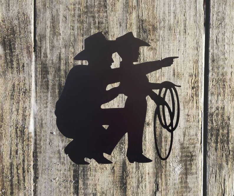 Puede incluir: Silueta met&aacute;lica negra de dos vaqueros, uno arrodillado y el otro se&ntilde;alando, con un lazo. La obra de arte est&aacute; montada sobre un fondo de madera envejecida, evocando un tema r&uacute;stico y del Oeste.