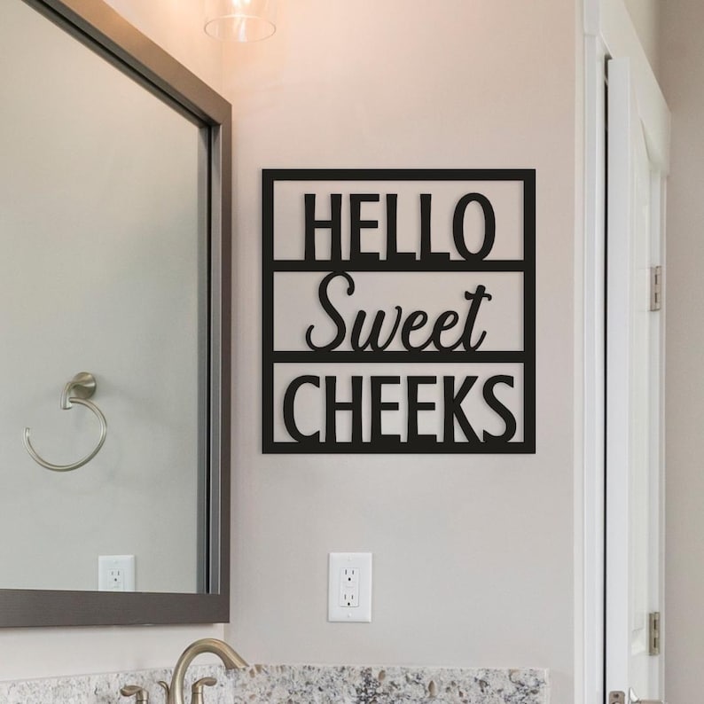 May include: Black metal wall art with the words "HELLO Sweet CHEEKS" in a three-sectioned rectangular frame. The sign is mounted on a light gray wall in a bathroom setting, near a mirror and a light switch.