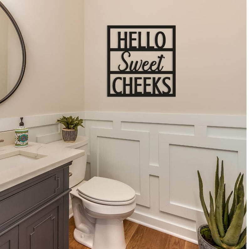 May include: Black metal wall art with the words "HELLO Sweet CHEEKS" in a bathroom setting. The sign is mounted on a white wall above a toilet. The bathroom has a neutral color scheme with a gray vanity and a round mirror.