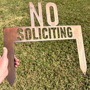 May include: A rusty metal sign with the words "NO SOLICITING" in large, bold letters. The sign is shaped like a rectangle with a pointed bottom.