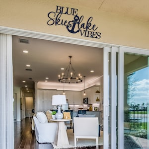 May include: A black metal sign with the text "BLUE SKIES and LAKE VIBES" and an anchor symbol. The sign is mounted on a beige wall above a sliding glass door leading to a patio.