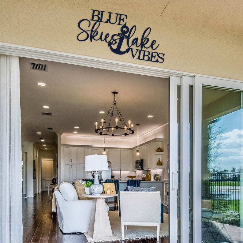 May include: A black metal sign with the text "BLUE SKIES and LAKE VIBES" and an anchor symbol. The sign is mounted on a beige wall above a sliding glass door leading to a patio.