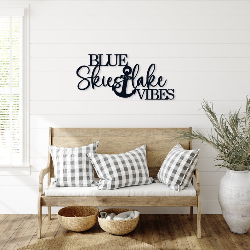 A black metal sign with the text 'BLUE SKIES AND LAKE VIBES' with an anchor symbol in between the words 'SKIES' and 'LAKE'. The sign is mounted on a white wall above a wooden bench with two checkered pillows.