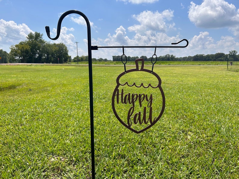 May include: A metal garden stake with a decorative acorn-shaped sign that reads "Happy Fall" in a cursive font. The sign is brown and hangs from a black metal hook. The background is a grassy field under a blue sky.