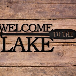 May include: Black metal sign with the words "Welcome to the Lake" and a stylized paddle design.