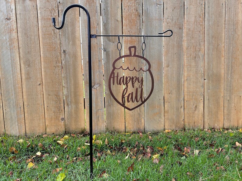 May include: A black metal garden stake with a decorative acorn-shaped sign that reads "Happy Fall" in a cursive font. The sign is made of brown metal and hangs from a horizontal bar. The background is a wooden fence.