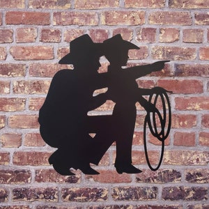 Puede incluir: Silueta met&aacute;lica negra de dos vaqueros, uno se&ntilde;alando, el otro en cuclillas. Los vaqueros llevan sombreros y sostienen un lazo. El fondo es una pared de ladrillo rojo.