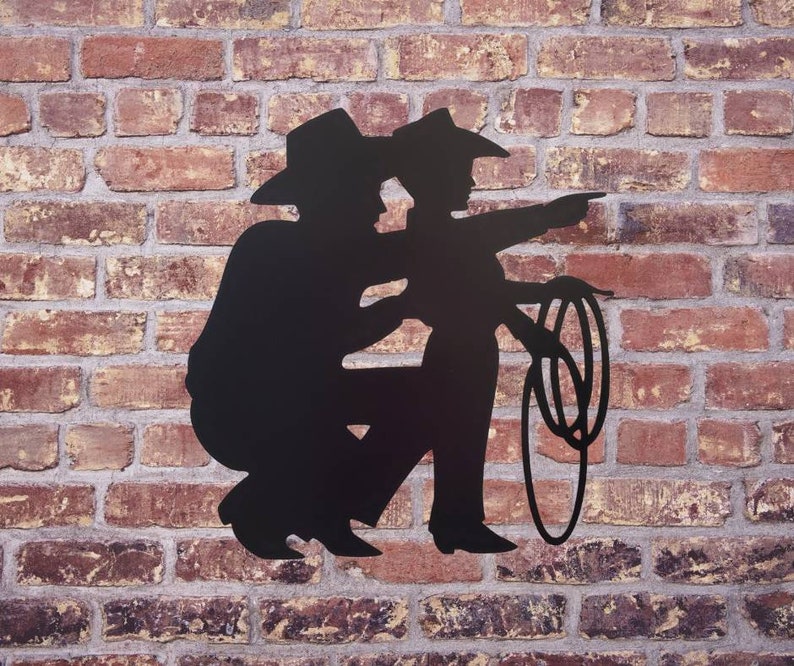 Puede incluir: Silueta met&aacute;lica negra de dos vaqueros, uno se&ntilde;alando, el otro en cuclillas. Los vaqueros llevan sombreros y sostienen un lazo. El fondo es una pared de ladrillo rojo.