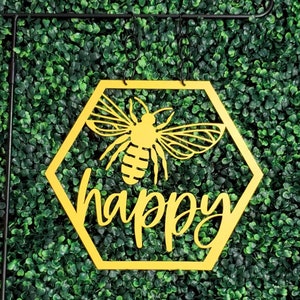 May include: A yellow hexagon-shaped metal sign with a cutout of a bee and the word "happy". The sign is hanging from a black metal hook.