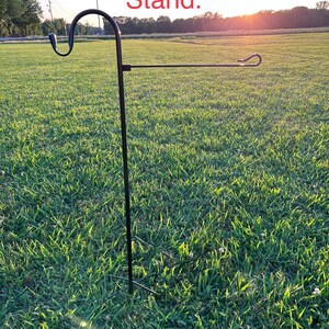 May include: A black metal garden flag stand with a curved top and a horizontal bar for displaying flags. The stand is set in a grassy field with the text "Premium Garden Flag Stand" in red at the top.
