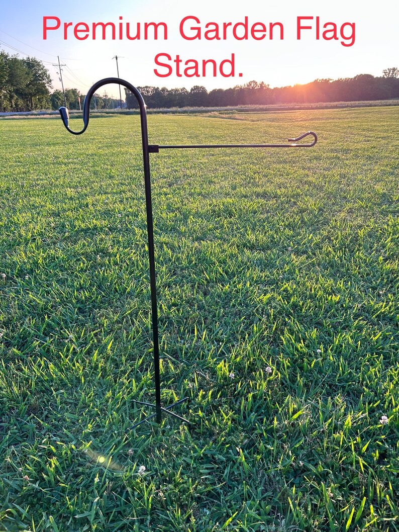 May include: A black metal garden flag stand with a curved top and a horizontal bar for displaying flags. The stand is set in a grassy field with the text "Premium Garden Flag Stand" in red at the top.