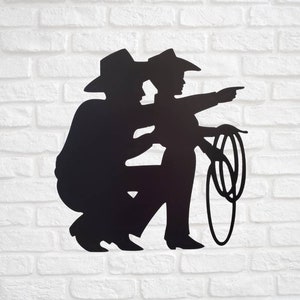 Puede incluir: Silueta met&aacute;lica negra de dos vaqueros, uno en cuclillas y el otro se&ntilde;alando con un lazo en la mano. Ambos llevan sombreros y botas de vaquero. La obra de arte est&aacute; montada en una pared de ladrillos blancos.