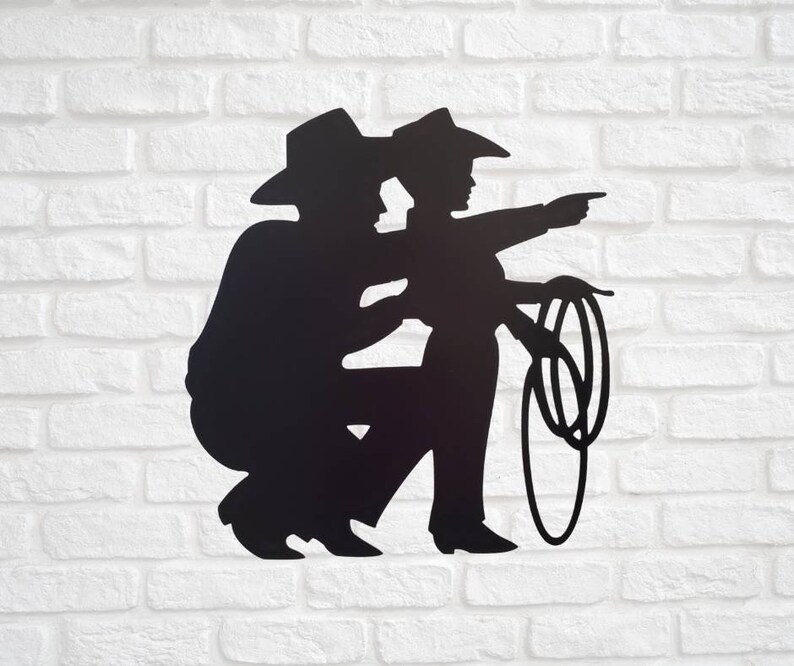 Puede incluir: Silueta met&aacute;lica negra de dos vaqueros, uno en cuclillas y el otro se&ntilde;alando con un lazo en la mano. Ambos llevan sombreros y botas de vaquero. La obra de arte est&aacute; montada en una pared de ladrillos blancos.