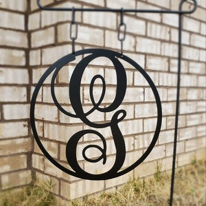 May include: Black metal garden flag with a circle design featuring a large, stylized letter G. The flag is hanging from a black metal stand against a brick wall.
