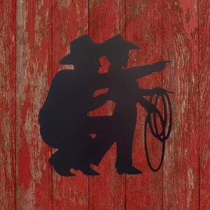 Puede incluir: Silueta met&aacute;lica negra de dos vaqueros, uno se&ntilde;alando, el otro sosteniendo un lazo. Las figuras se alzan sobre un fondo de madera roja desgastada, evocando un tema r&uacute;stico y occidental.