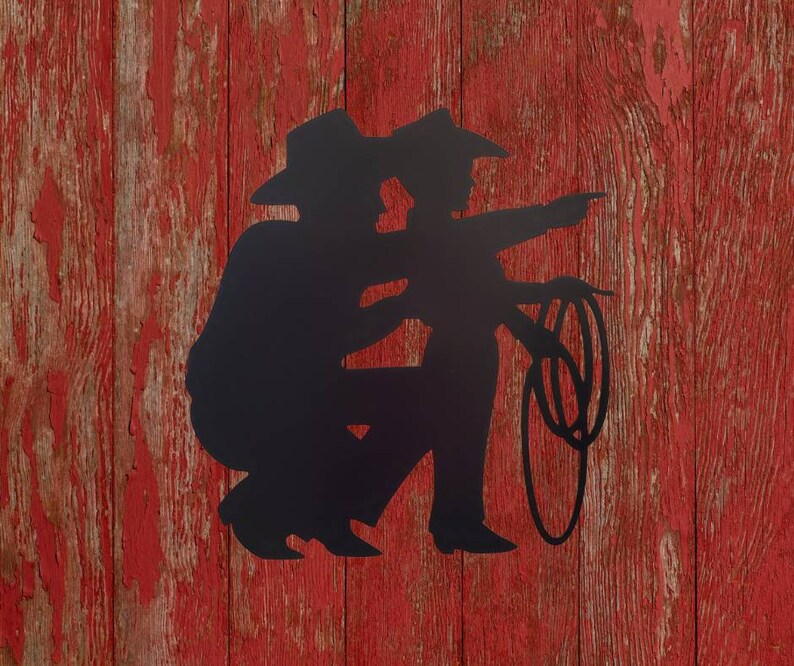 Puede incluir: Silueta met&aacute;lica negra de dos vaqueros, uno se&ntilde;alando, el otro sosteniendo un lazo. Las figuras se alzan sobre un fondo de madera roja desgastada, evocando un tema r&uacute;stico y occidental.