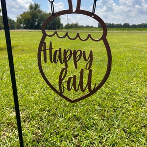 May include: A brown metal acorn-shaped sign with the words "Happy Fall" cut out. The sign is hanging from a black chain against a backdrop of green grass and a blue sky.
