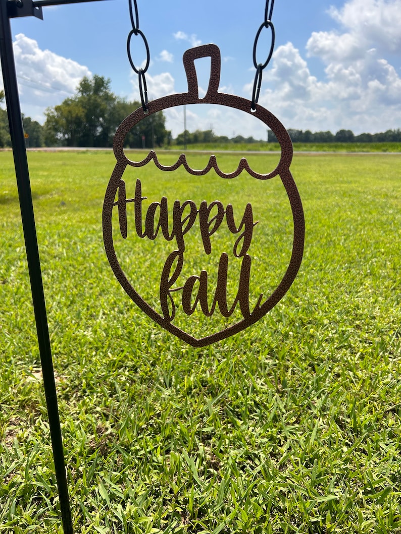 May include: A brown metal acorn-shaped sign with the words "Happy Fall" cut out. The sign is hanging from a black chain against a backdrop of green grass and a blue sky.