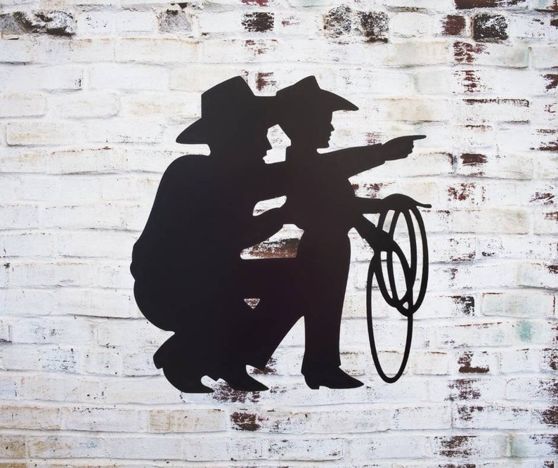 Puede incluir: Silueta met&aacute;lica negra de dos vaqueros, uno se&ntilde;alando y el otro sosteniendo un lazo. Las figuras se colocan contra una pared de ladrillos blancos, creando un dise&ntilde;o r&uacute;stico y tem&aacute;tico del oeste.
