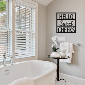 May include: A black metal wall sign in a bathroom with the words "HELLO Sweet CHEEKS". The sign is mounted on a light gray wall above a small table with a glass jar, towels, and a potted plant. A white bathtub is in the foreground.