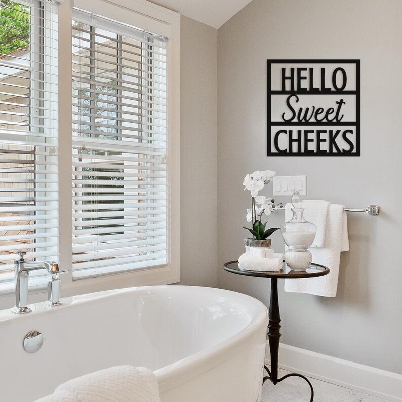 May include: A black metal wall sign in a bathroom with the words "HELLO Sweet CHEEKS". The sign is mounted on a light gray wall above a small table with a glass jar, towels, and a potted plant. A white bathtub is in the foreground.