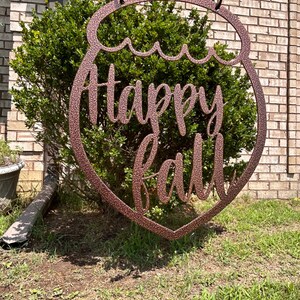 May include: A copper-colored metal sign with the words "Happy Fall" in a cursive font. The sign is shaped like a decorative ornament and hangs from black chains. The sign is displayed outdoors in front of a green bush and brick wall.