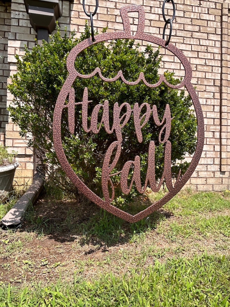 May include: A copper-colored metal sign with the words "Happy Fall" in a cursive font. The sign is shaped like a decorative ornament and hangs from black chains. The sign is displayed outdoors in front of a green bush and brick wall.