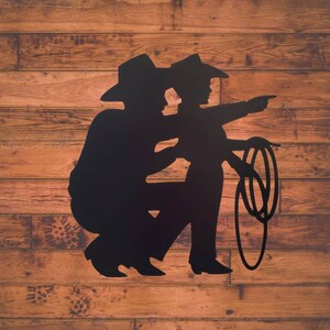 Puede incluir: Silueta negra de dos vaqueros, uno arrodillado y el otro se&ntilde;alando, con un lazo. La obra est&aacute; sobre un fondo de tablones de madera, creando una est&eacute;tica r&uacute;stica.
