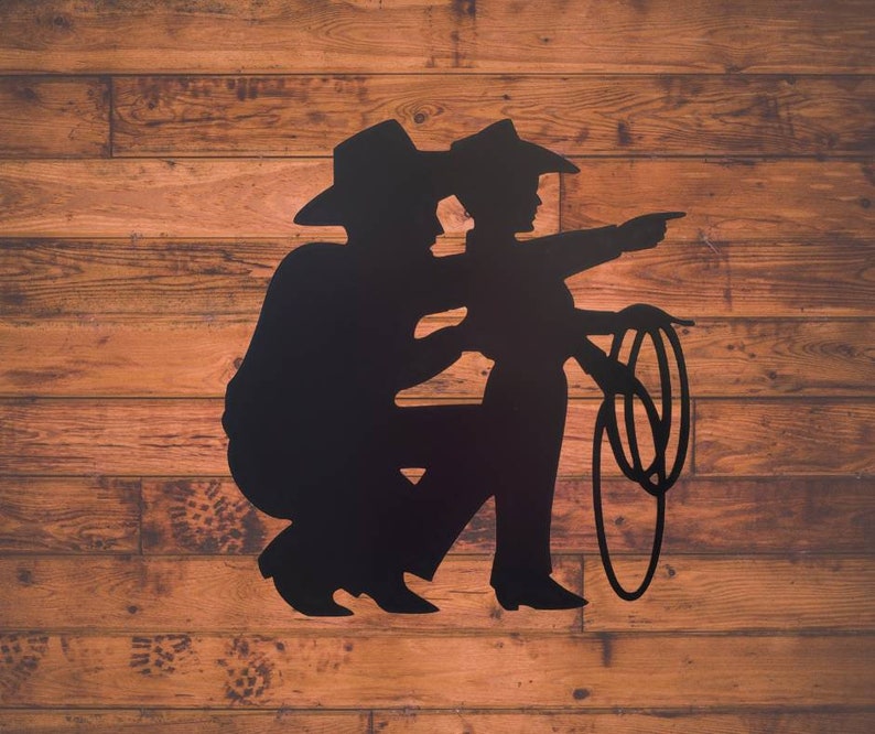 Puede incluir: Silueta negra de dos vaqueros, uno arrodillado y el otro se&ntilde;alando, con un lazo. La obra est&aacute; sobre un fondo de tablones de madera, creando una est&eacute;tica r&uacute;stica.