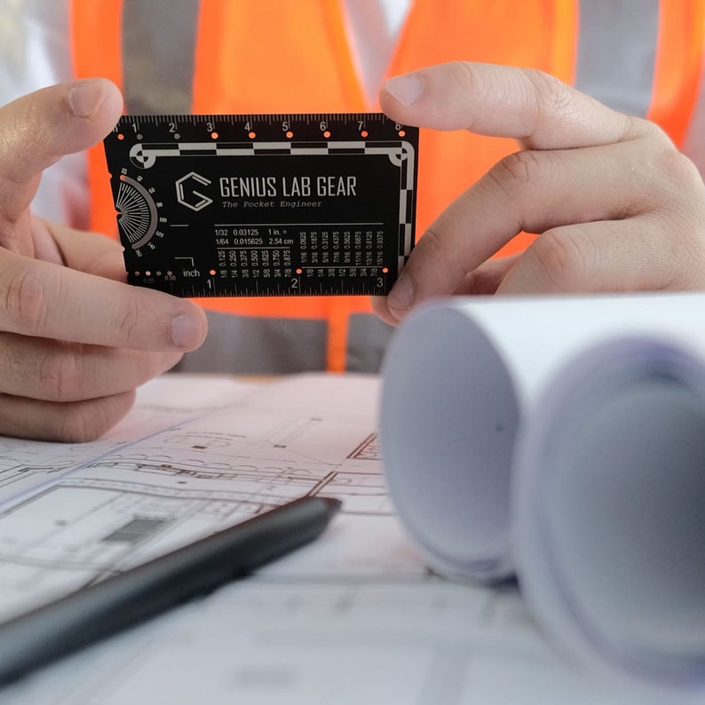 May include: A black metal ruler with the text "GENIUS LAB GEAR The Pocket Engineer" and measurements in inches and centimeters. The ruler is being held by a person's hands.