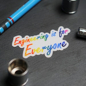 May include: A white sticker with the text "Engineering is for Everyone" in rainbow colors. The sticker is on a dark gray surface.