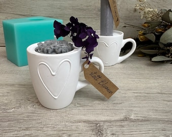 May include: Two white ceramic mugs with heart-shaped handles, one with a gray candle inside and the other with a small gray candle holder. Both mugs have a tag attached, one says "statt Blumen" and the other says "statt Blumen".