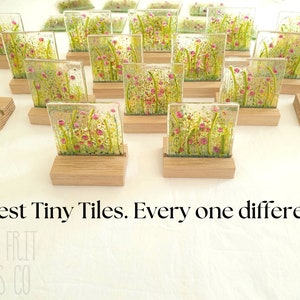 May include: A collection of small, square glass tiles with a floral design. Each tile is unique and features a different arrangement of pink flowers and green foliage. The tiles are displayed on small wooden stands. "Latest Tiny Tiles. Every one different." is written below the tiles.