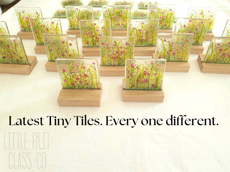 May include: A collection of small, square glass tiles with a floral design. Each tile is unique and features a different arrangement of pink flowers and green foliage. The tiles are displayed on small wooden stands. "Latest Tiny Tiles. Every one different." is written below the tiles.