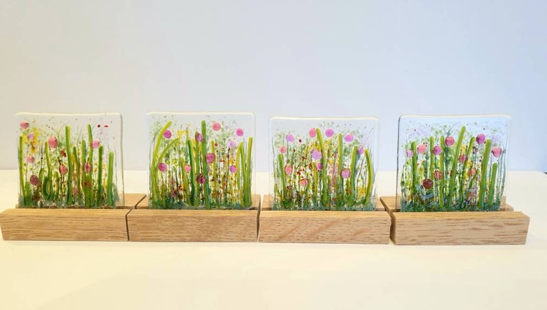 May include: Four square glass panels with a painted design of pink and yellow flowers in a green field. Each panel is mounted on a small wooden stand.