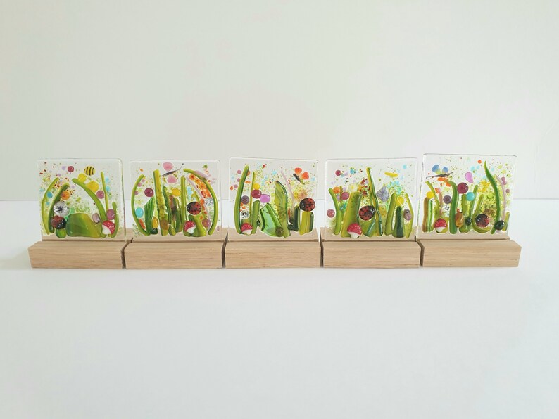May include: Five small glass squares with colorful images of a garden scene with flowers, mushrooms, ladybugs, and dragonflies. Each square is mounted on a small wooden base.