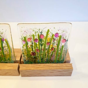 May include: Two square glass coasters with a floral design. The coasters feature a white background with green stems and pink flowers. The coasters are displayed on a wooden stand.