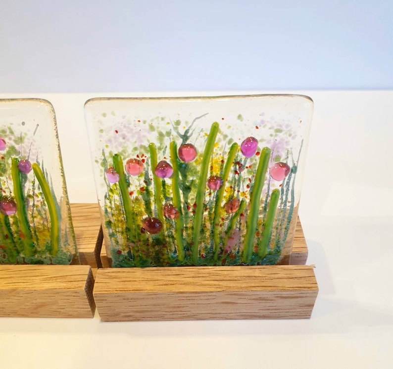 May include: Two square glass coasters with a floral design. The coasters feature a white background with green stems and pink flowers. The coasters are displayed on a wooden stand.