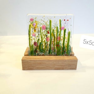 May include: A small glass art piece depicting a field of green grass with pink and red flowers. The piece is displayed on a wooden stand. 5x5cm.