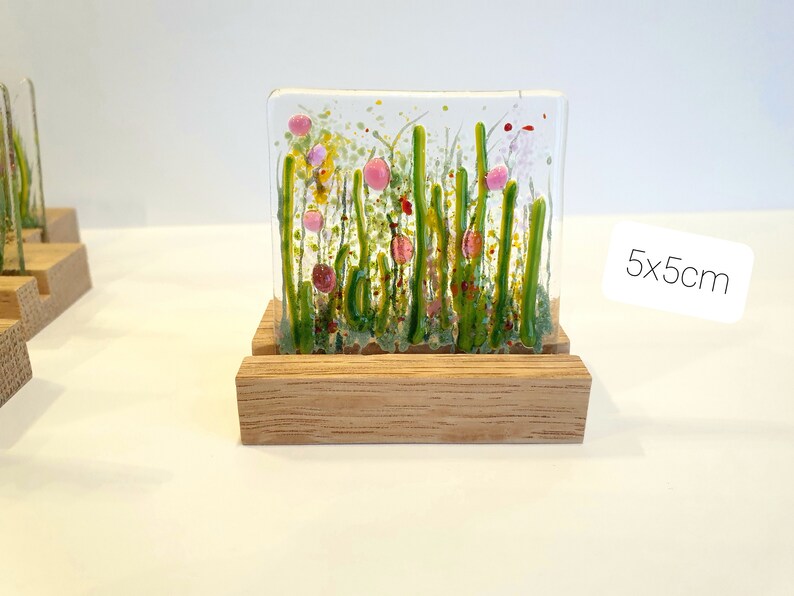 May include: A small glass art piece depicting a field of green grass with pink and red flowers. The piece is displayed on a wooden stand. 5x5cm.