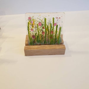 May include: A glass art piece depicting a field of green grass with pink and red flowers. The piece is displayed on a wooden stand.