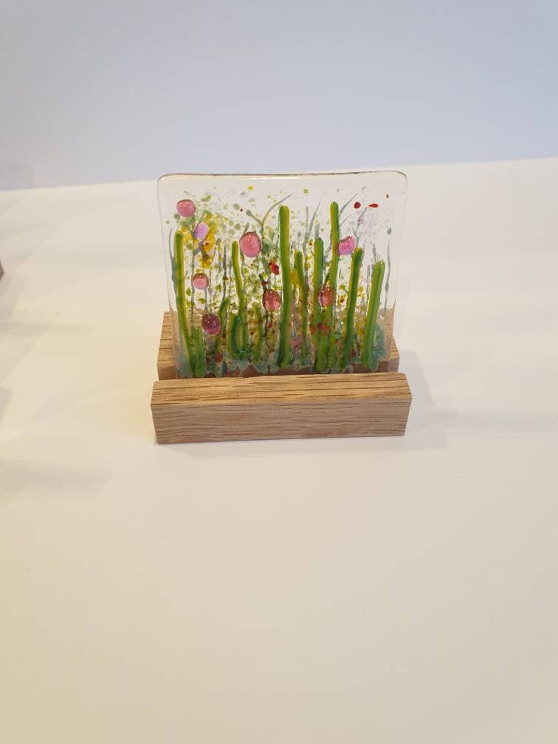 May include: A glass art piece depicting a field of green grass with pink and red flowers. The piece is displayed on a wooden stand.