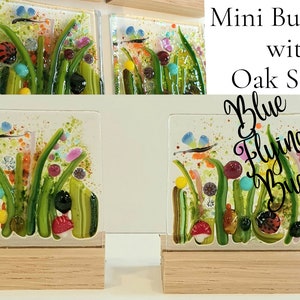 May include: Two small glass tiles with a colorful, abstract design of green grass, flowers, and blue flying bugs. The tiles are on wooden stands. The text "Blue Flying Bugs" is written on the image.