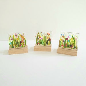 May include: Three glass art pieces on wooden stands, each depicting a colorful, abstract floral design with butterflies. The glass is clear and the flowers are in shades of pink, green, yellow, and blue.