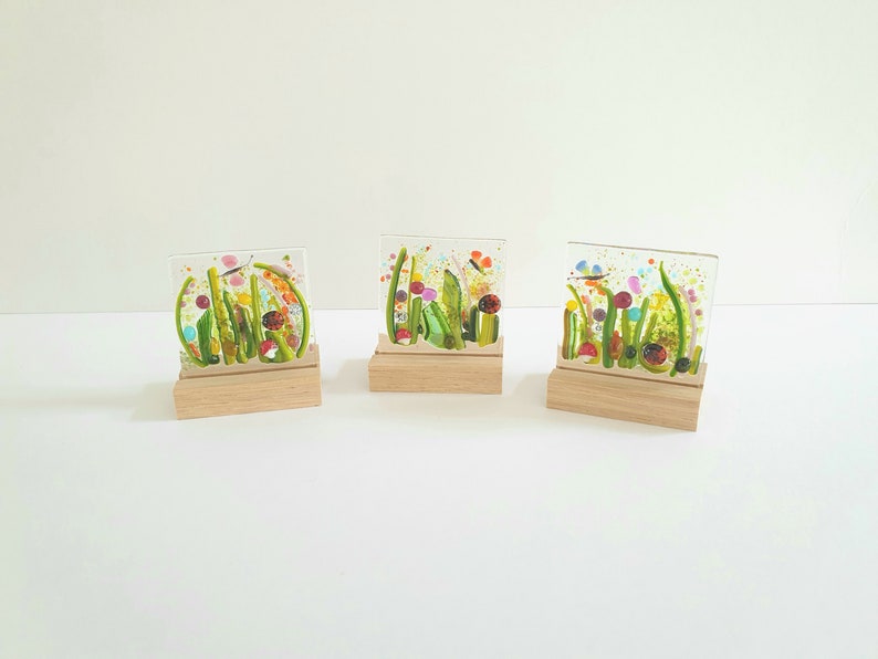 May include: Three glass art pieces on wooden stands, each depicting a colorful, abstract floral design with butterflies. The glass is clear and the flowers are in shades of pink, green, yellow, and blue.