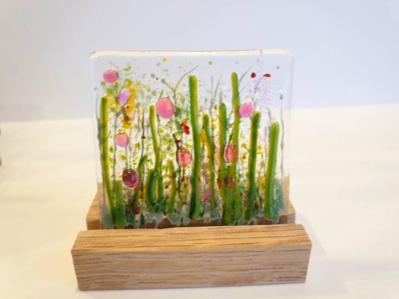 May include: A glass art piece depicting a field of green grass with pink flowers. The piece is displayed on a wooden stand.
