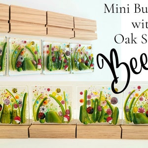 May include: Five small glass tiles with a nature scene of green grass, flowers, mushrooms, and bees. The tiles are displayed on wooden stands. The text "Mini Bug Tiles with Oak Stands Bees" is written above the tiles.