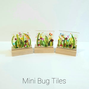 May include: Three small glass tiles with a nature scene of green grass, flowers, and butterflies. The tiles are displayed on small wooden stands. The text "Mini Bug Tiles" is printed below the tiles.