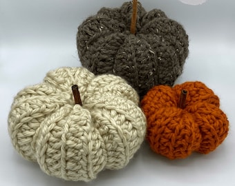 May include: Three crocheted pumpkins in varying sizes and colors. The largest is a speckled brown, the medium is cream, and the smallest is orange. Each pumpkin has a cinnamon stick stem. The image includes the text "Huff's Crochet Stuff Handmade Gifts."
