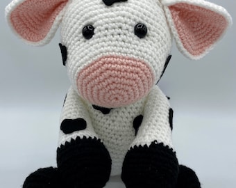 May include: A handmade crochet cow plushie. The cow is primarily white with black spots, black hooves, and a pink muzzle and inner ears. It has small, tan horns and large black eyes. The plushie is sitting upright. The image includes the text "Huff's Crochet Stuff Handmade Gifts".